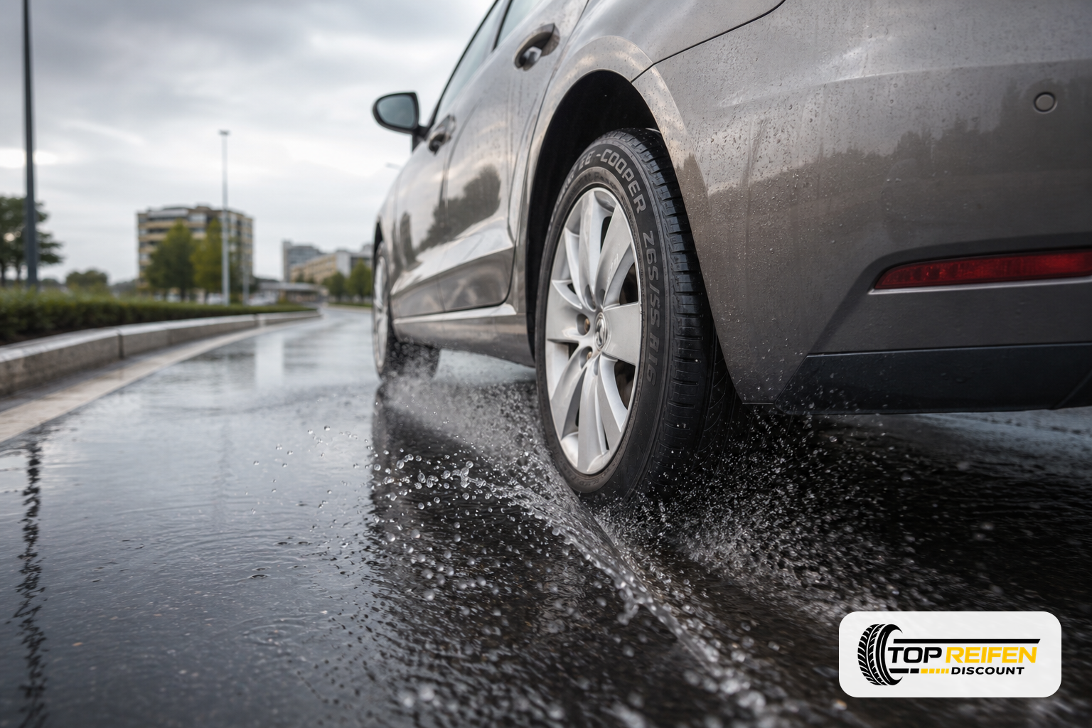 COOPER ZE-CS8 205/55 R16 auf nasser Stadtstraße: hinteres Rad eines PKW wirft feinen Sprühnebel auf dunklem Asphalt bei bedecktem Himmel.